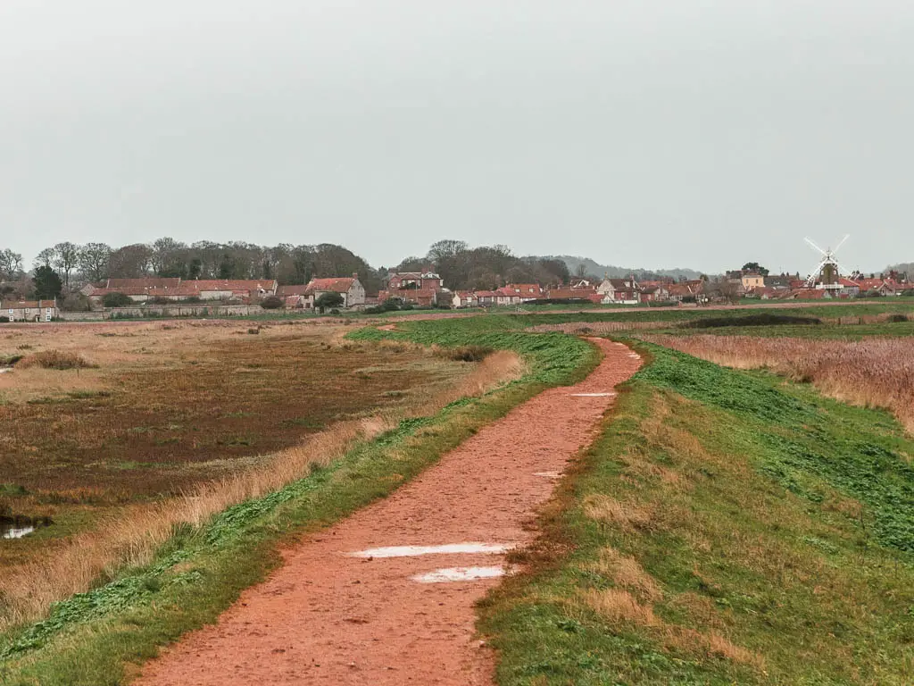 A ridgeway path leading towards a village. The path is lined with grass and marshland on either side.