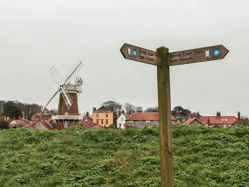 A wooden trail signpost marking the walk along the Norfolk Coastal Path. There is a green grass hill behind the sign with village houses and a windmill behind.