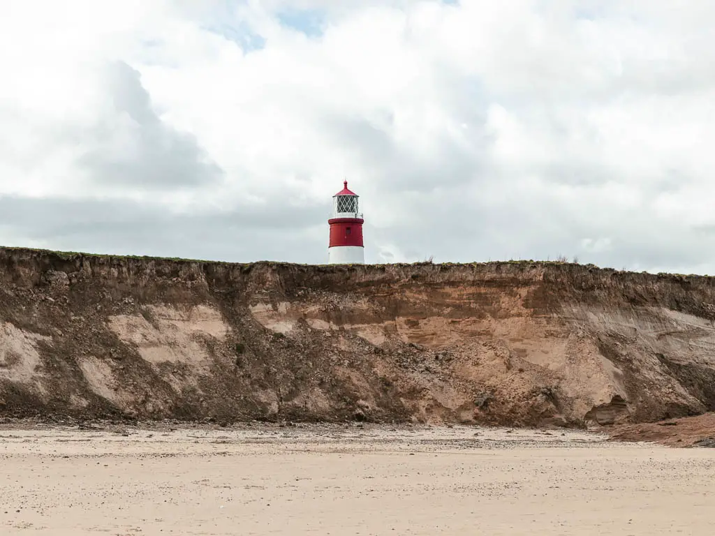 Looking across the beach to the rugged cliff and a red and white stripped lighthouse on top, on the walk along the Norfolk Coast Path.