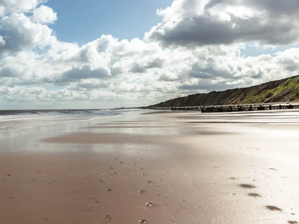 A long wide empty sandy beach when walking the coast path in Norfolk. There are grass covered cliffs to the right, and the sea to the left.