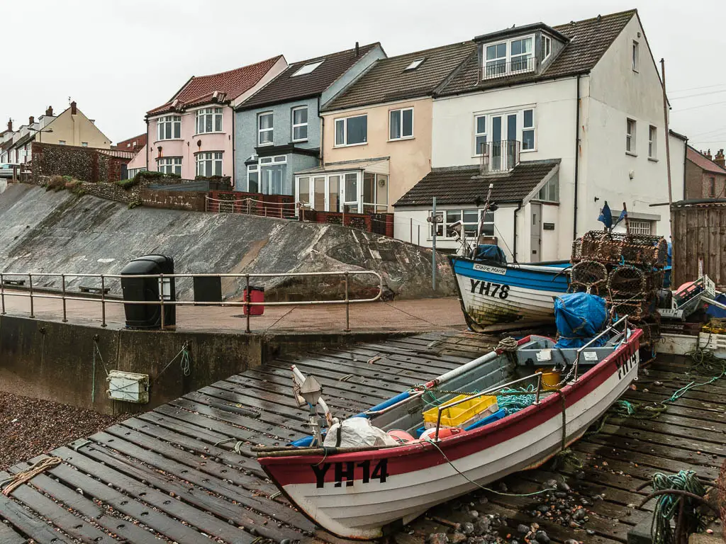 Small boats with fishing equipment on the sea ramp, with a walking path and village houses behind them up a hill.