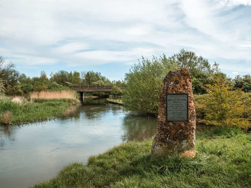 A stone plaque on the grass bank, with the river on the left and a bridge over the river ahead.