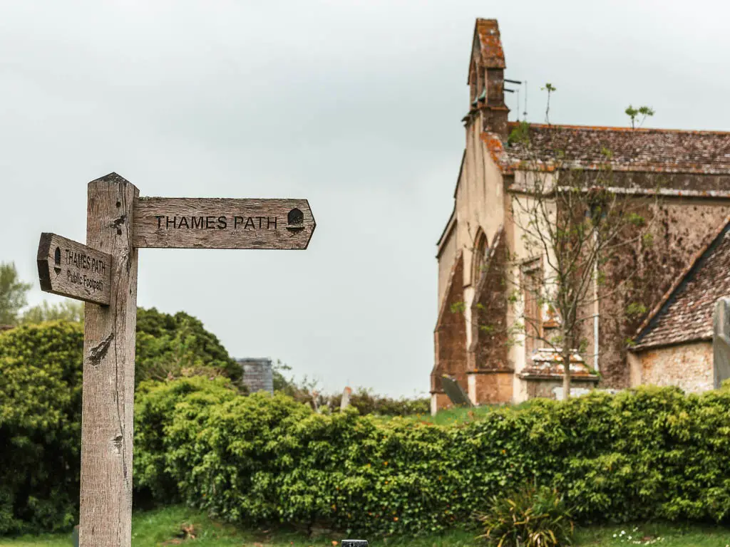 A wooden trail sign on the left and church ahead, on the walk towards Lechlade from Cricklade. The sign points right and back.
