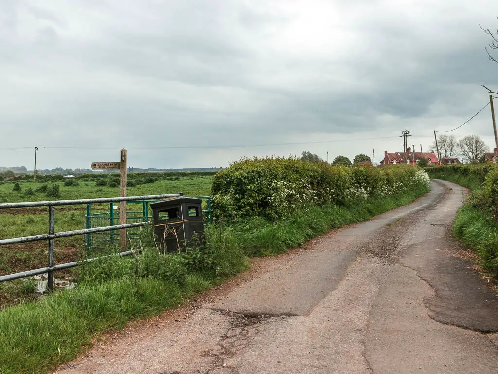 A road curving ahead on the right, with a green grass field on the left. The field is lined with a metal fence and green hedges.