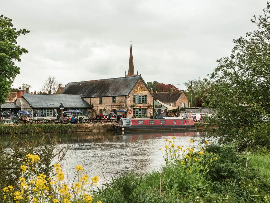 Looking over the grass and yellow flowers to the river, and the Riverside pub on the other side at the end of the walk from Cricklade to Lechlade. There is a barge moored to the episode of the river.