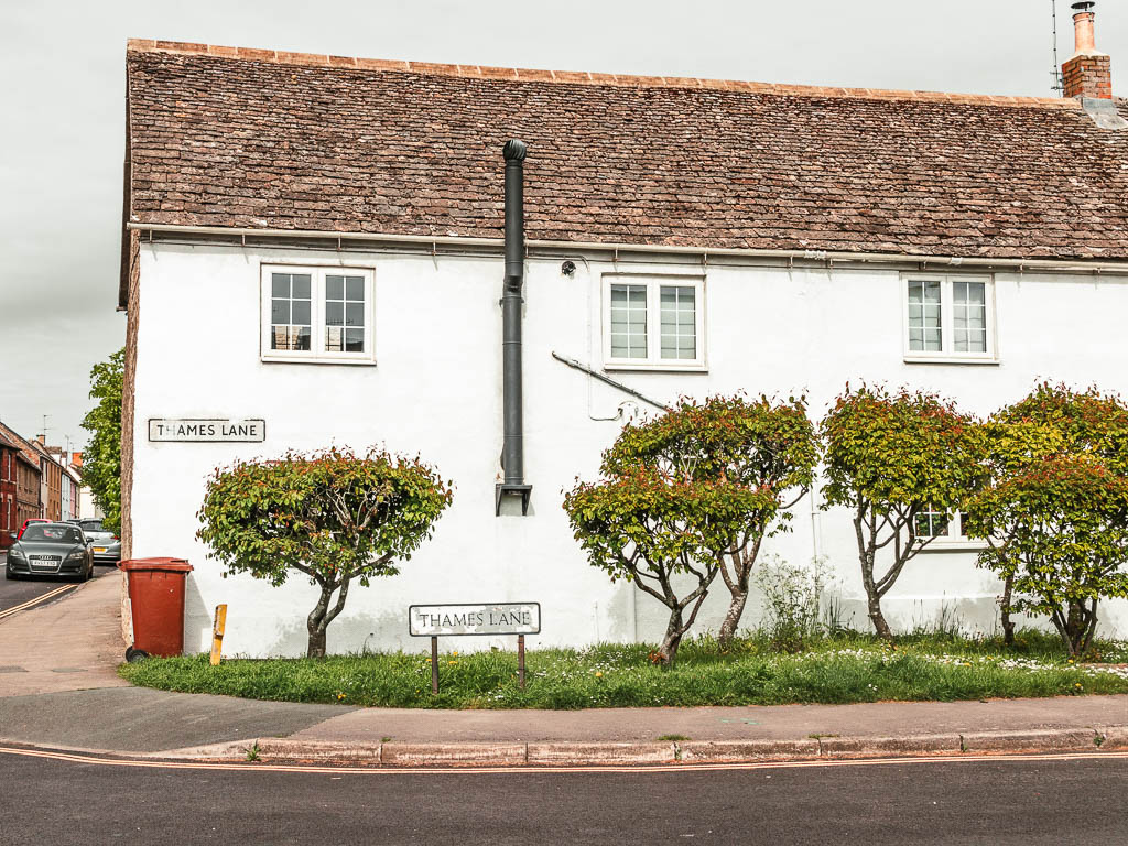 A white walled house on the4 road corner, with a sign saying 'thames Lane' There are small trees in front of the house.