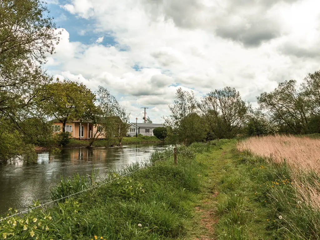 The river on the left leading ahead, with strip of grass on the right side, and partially hidden buildings of the holiday park on the other side.