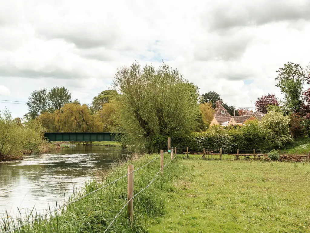 A grass field on the right, and river on the left and a wire fence separating the two. There are trees ahead, and rooftops of houses visible.