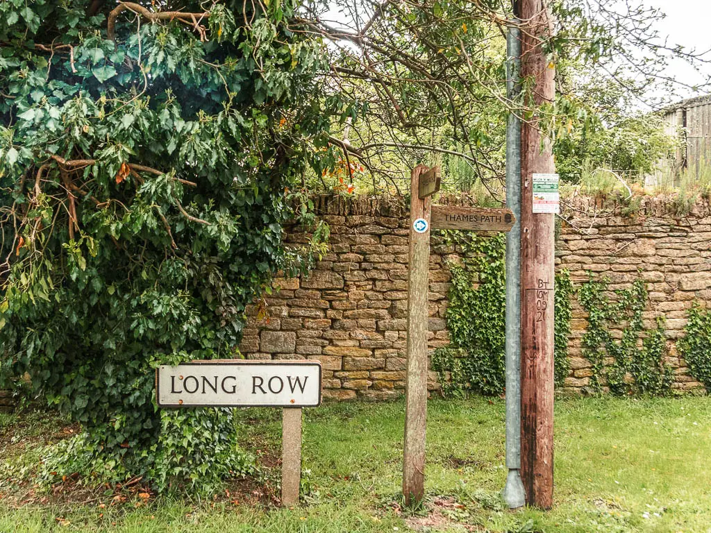 A road sign saying 'Long Row' on the grass bank, with a stone all and leafy bushes behind it, and a wooden trail sign on the right.