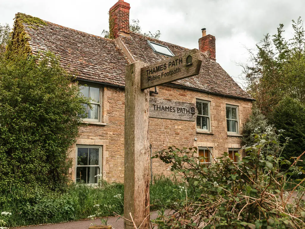 A wooden trail signpost saying 'Thames Path public footpath' sitting in front of a stone walled house on the walk between Cricklade and Lechlade. The sign points right and back.