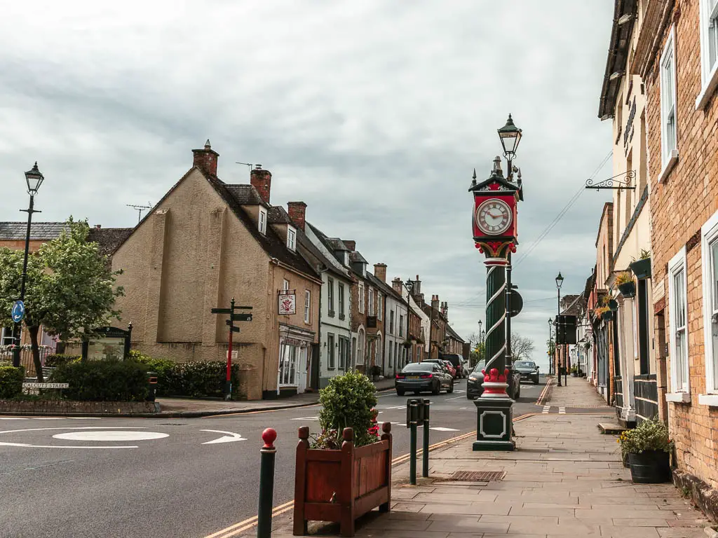 A road and roundabout on the left and pavement with tall clock on the right in Cricklade. The road is lined with buildings on both side.