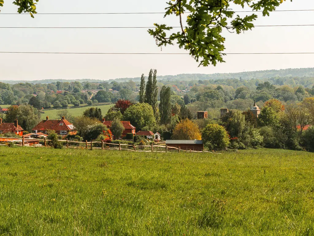 Looking down a large grass field to village buildings below, and a vast view to a tree covered landscape in the distance.