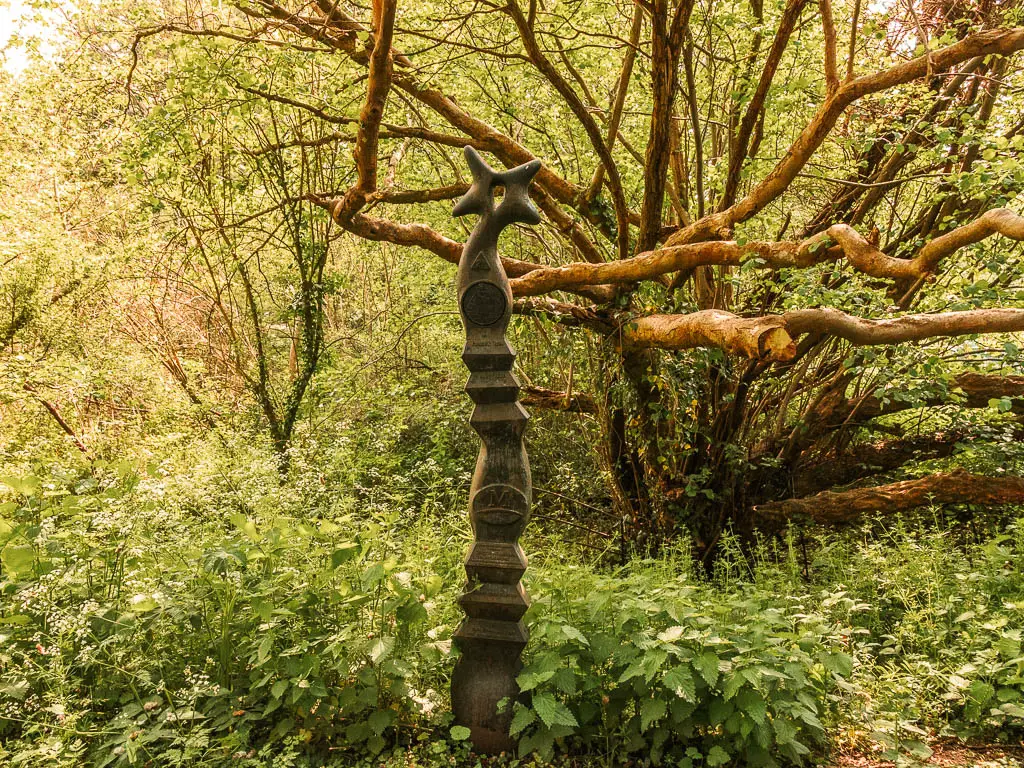 A sculpture in front of green leafy bushes and a tree with lots of straggly branches.