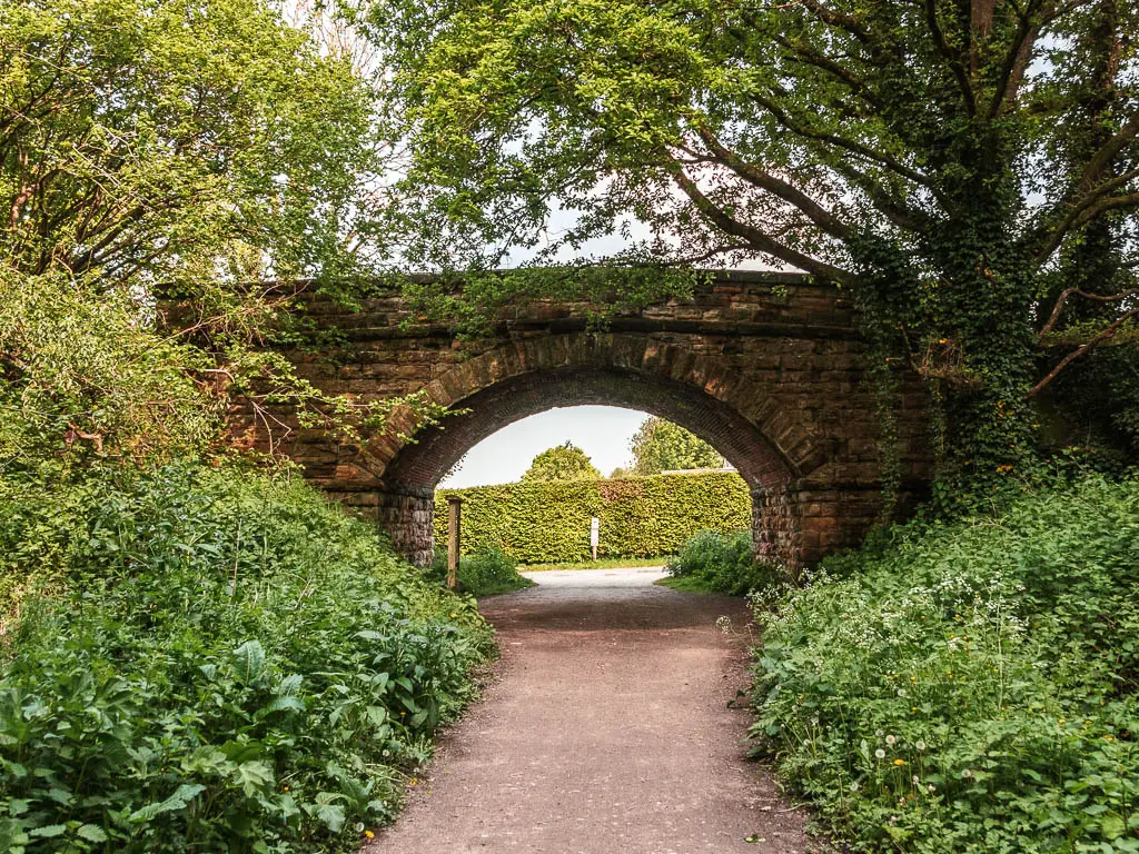 A wide path leading under and archway bridge made of stone. Through the bridge there is a neatly cut hedge lining the road.