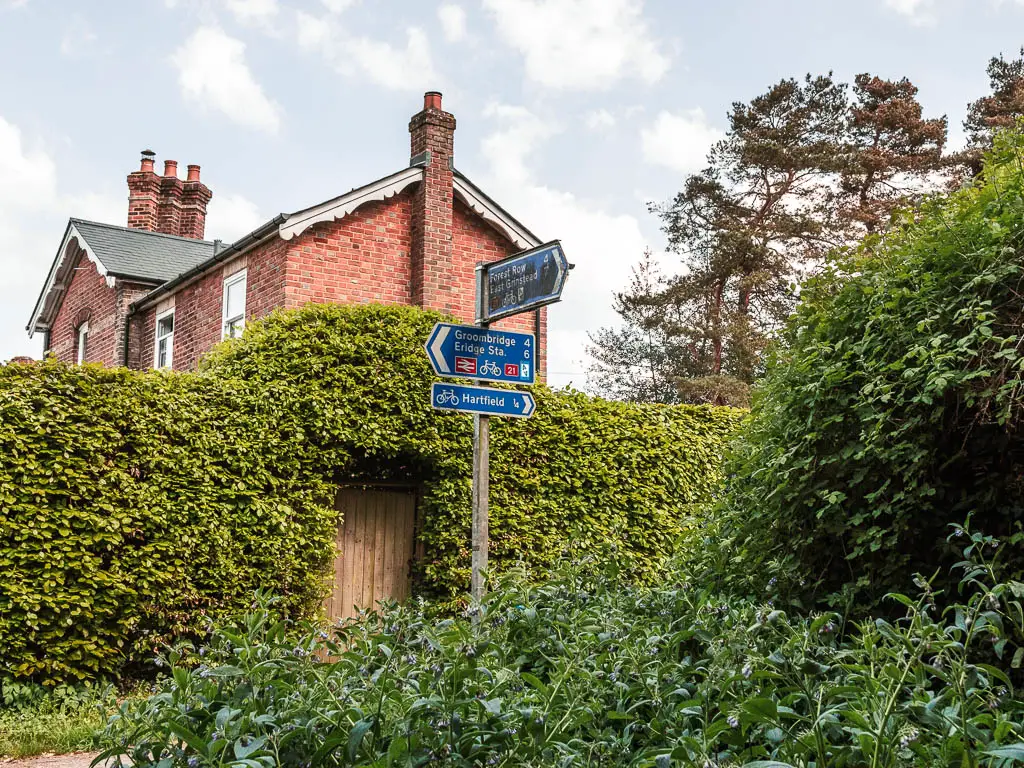 A blue trail sign post coming out of the bushes, pointing left to walk to Groombridge along the Forest Way. There his a hedge behind the trail and there top half of a brick house visible on the other side.