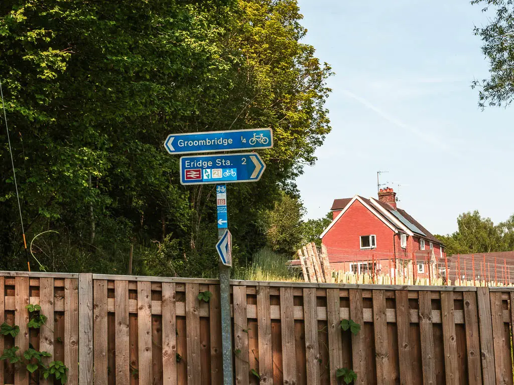 A blue signpost in front of a wooden fence, pointing left to walk to Groombridge.  There is a big leafy green tree behind the sign, and a red walled house in the distance on the right.