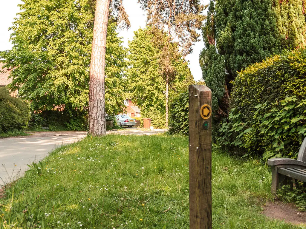 A wooden stump sign post on the grass bank, with the road on the left. the arrow on the sign points right.