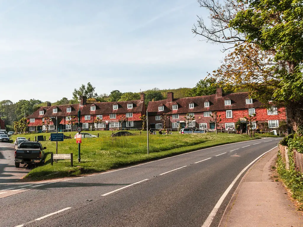 A road leading uphill from left to right, and a green ahead to the left, with quaint houses lined up ahead along the top of the green.