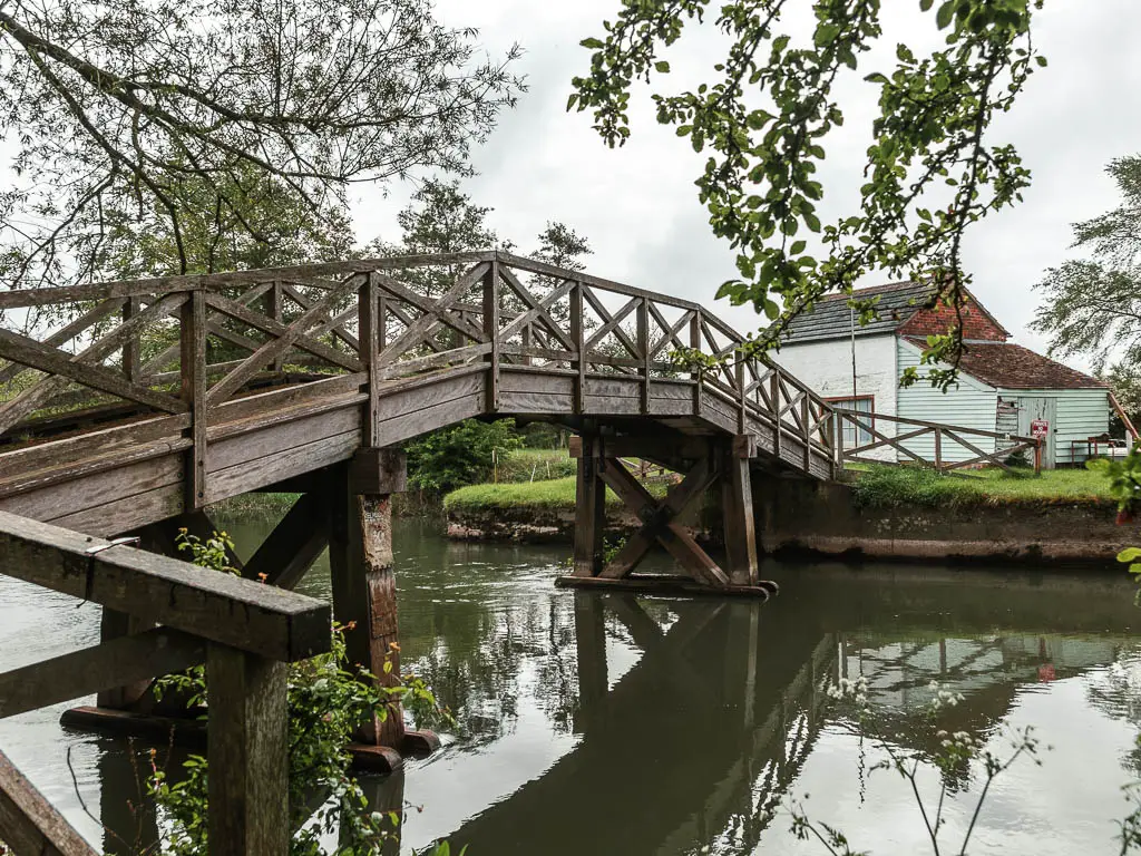 A wooden bridge over the river, and white walled house on the other side, partway through the walk from Lechlade to Newbridge. There are tree branches with green leaves hanging in the frame.