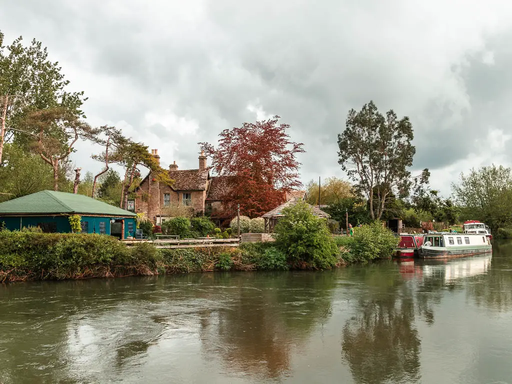 Looking across the river to the other side with bushes and buildings surround with red and green leafed trees, part way through the walk from Lechlade to Newbridge. There are two barges moored to the other side.