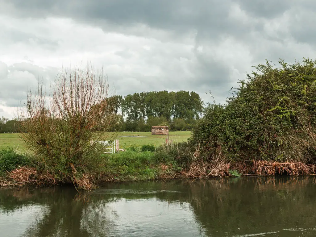 Looking across the river to a large green grass field and pillbox in the middle of it.