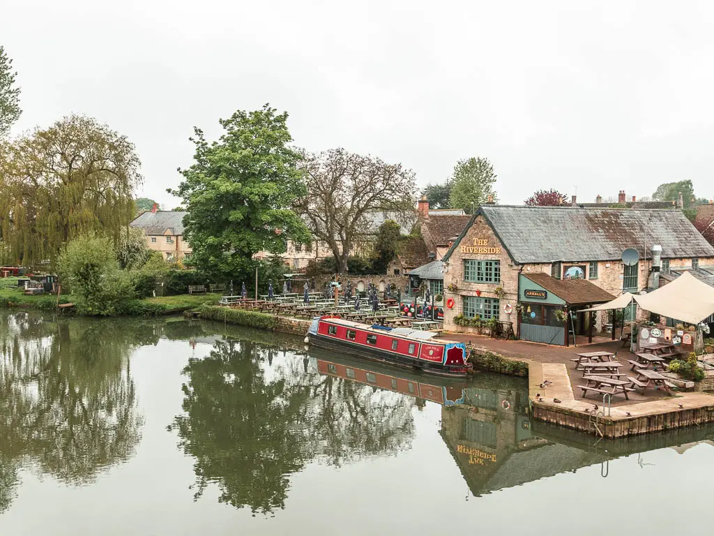 Looking down to the river with the Riverside pub on the right side, in Lechlade. There is a red barge moored to the side.