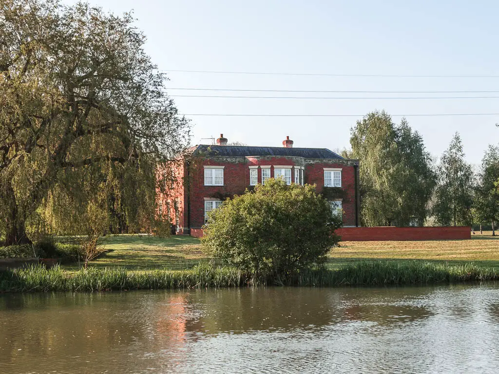 Looking across the river the a neatly cut grass field on the other side and a large red brick walled manor house. There is a green leafy tree partially blocking the view of the house.