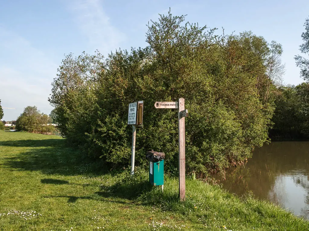 A wooden trial signpost on the green, with the river to the river. There is a big leafy tree behind the sign. 