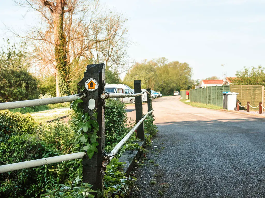 A gravel road leading ahead to the car park, and yellow acorn and yellow arrow trail sign on the post on the railing on the left.