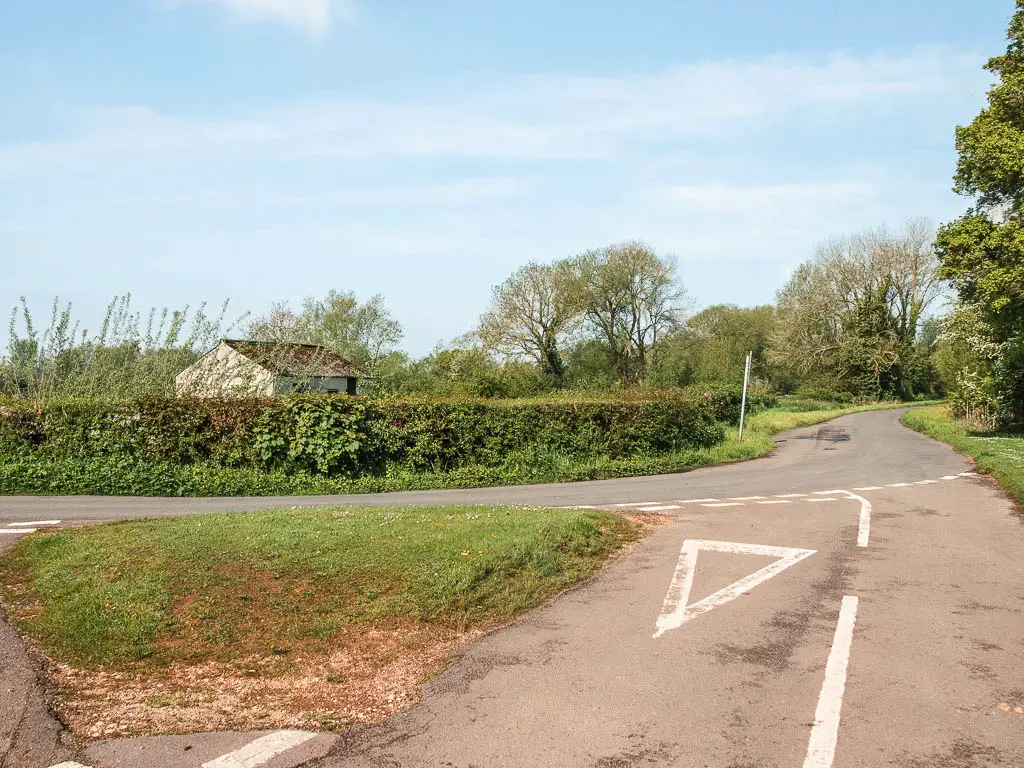 The road leading to the corner of the main road. The other side of the main road is lined with a green hedge and the rooftop of a bark poking out from the other side.