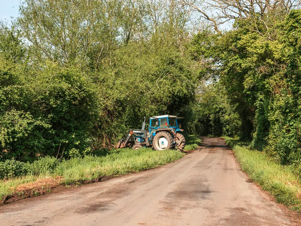 A road leading straight ahead, lined with strips of grass and green leafy bushes and trees on the walk from Newbridge to Oxford. There is a blue tractor parked on the left side.