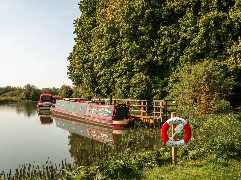 A barge moored to the side of the calm river with reflections, on the walk from Newbridge to Oxford. On the right side is grass, bushes and big green leafy trees. There is a hanging raft ring in white and red saying 'Pinkhill Lock'.