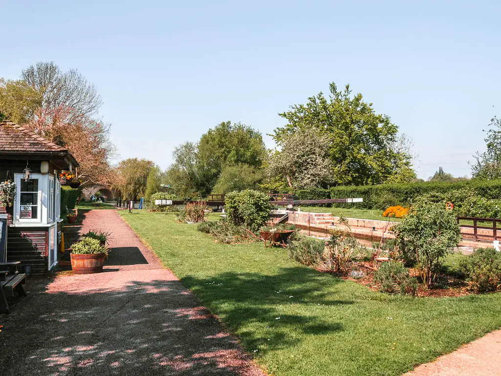 A path on the left and neatly cut green on the right, with planted flowers. There is a wooden hut just visible on the left side of the path.