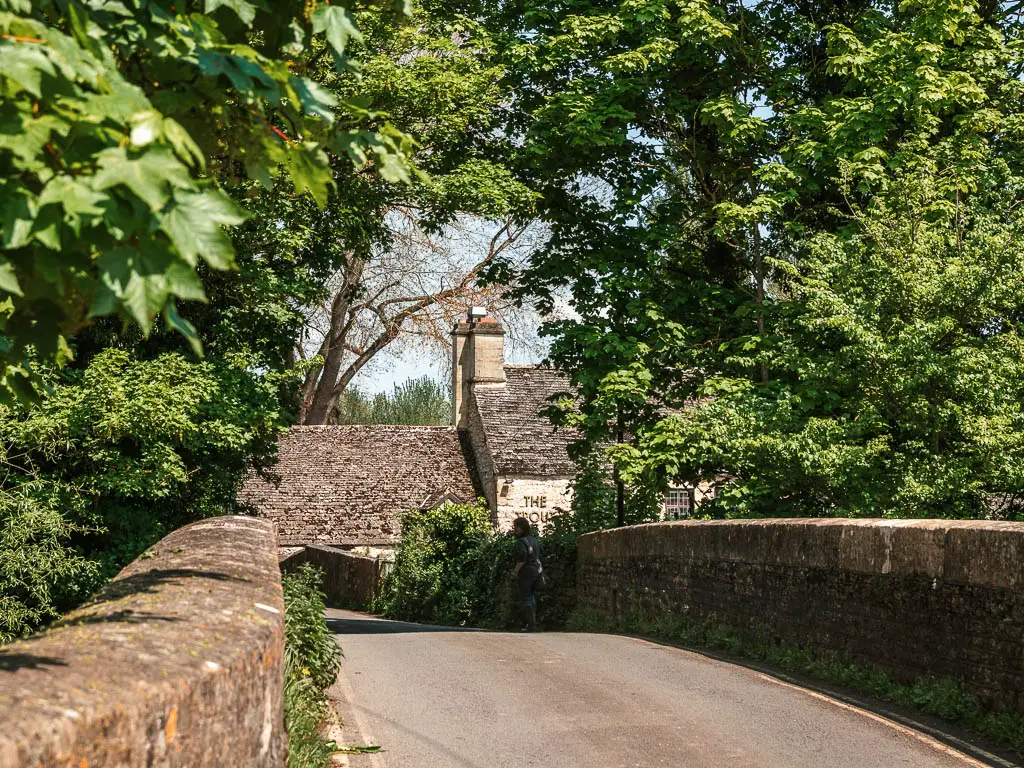 Looking along the road lined with stone walls and part of a pub visible on the other side. There are big green leafy trees on the other side of both stone walls, and over hanging.