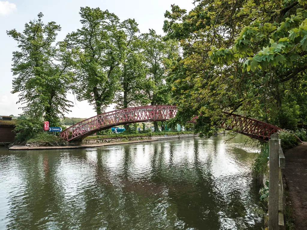 Looking along the river with ripples, towards a red metal brush. There are green leafy tree branches hanging over the bridge and river.