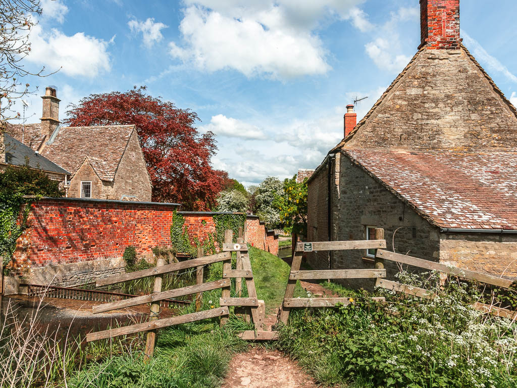 A wooden fence with a V gap and old stone village buildings ahead, partway through the walk from the source to Cricklade.