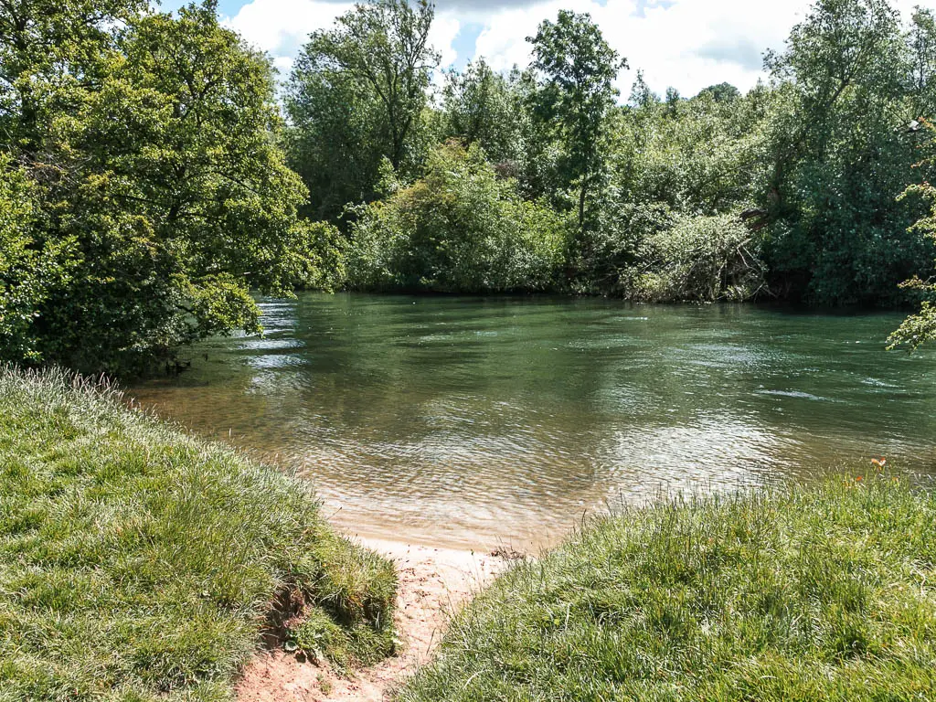 A small patch of white sand in front of the grass bank with the river with clear water, on the walk from Marlow to Maidenhead. The other side of the river is lined with trees.