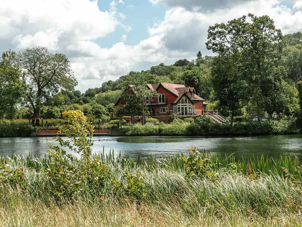 Looking over the tall grass to the river and a big house on the other side, when walking from Marlow to Maidenhead. The house is partially hidden by trees.