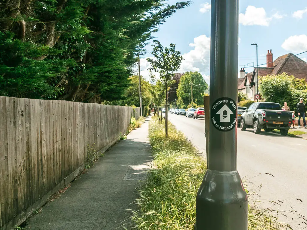 A metal light pole with thames path sticker pointing ahead with a white arrow. There is a pavement leading ahead with the road on the right and a Woden fence on the left. There are green leafy trees behind the fence. There are cars parked on the right side of the road.