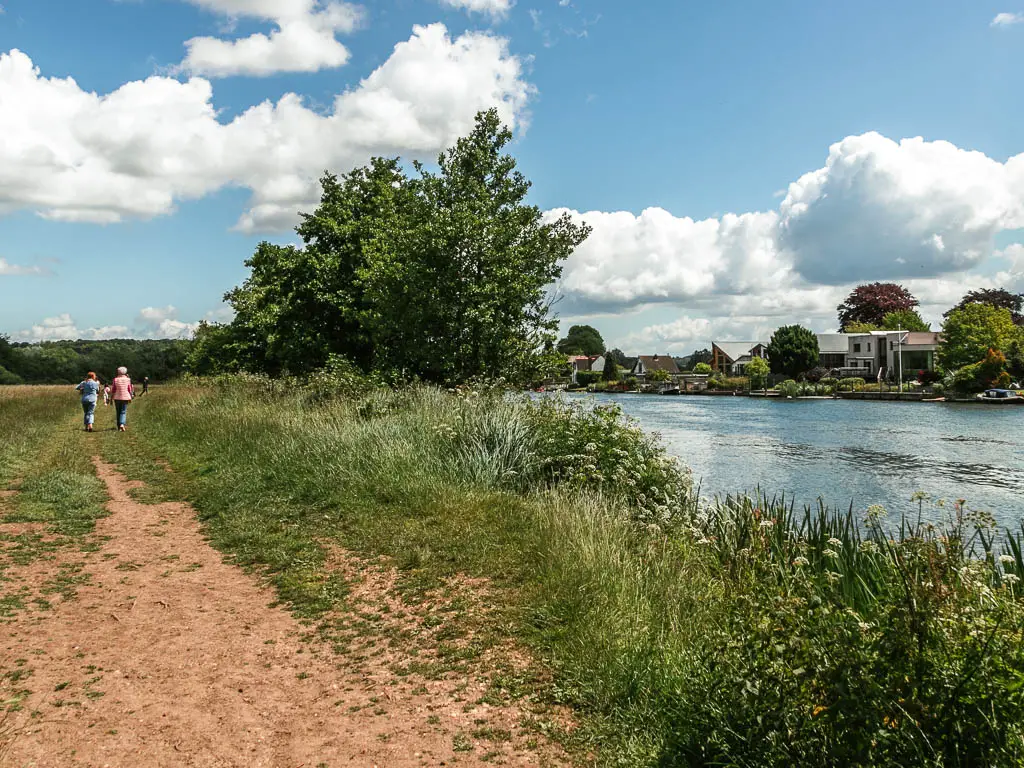 A dirt trail through the grass on the left with the river on the right. There is tall grass and bushes lining the river edge. There are two people walking along the trail ahead.