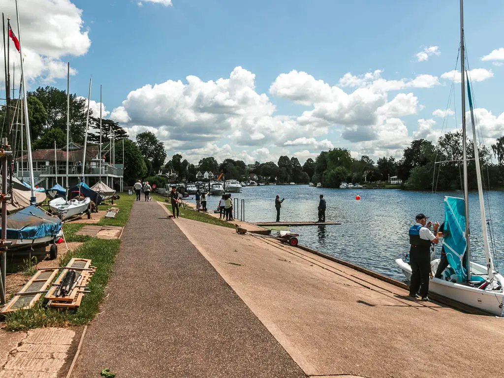 A path leading ahead with slope to the river on the right. There are sailboats on the ground on the left and one on the right in the river. There are a few people walking ahead.