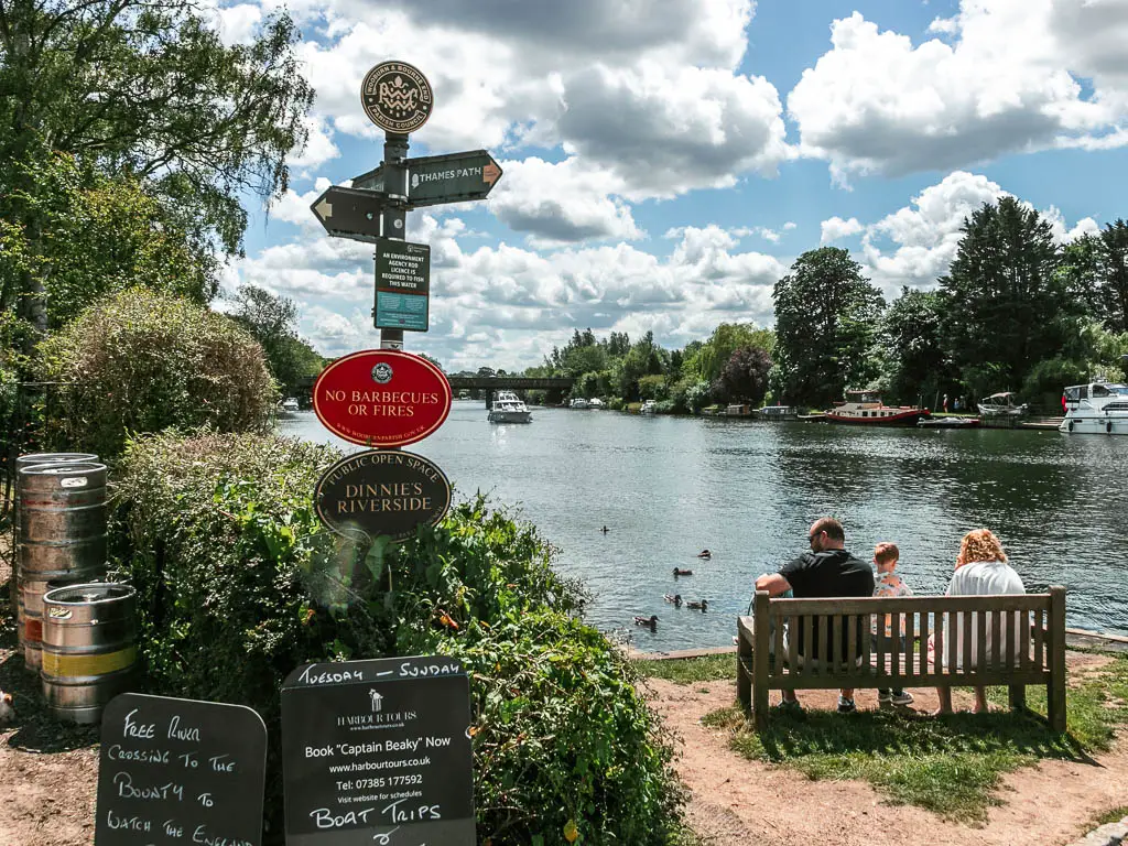 A signpost pointing in all directions in front of a hedge. There river is to the right. There is a wooden bench facing the river with three people sitting on it.