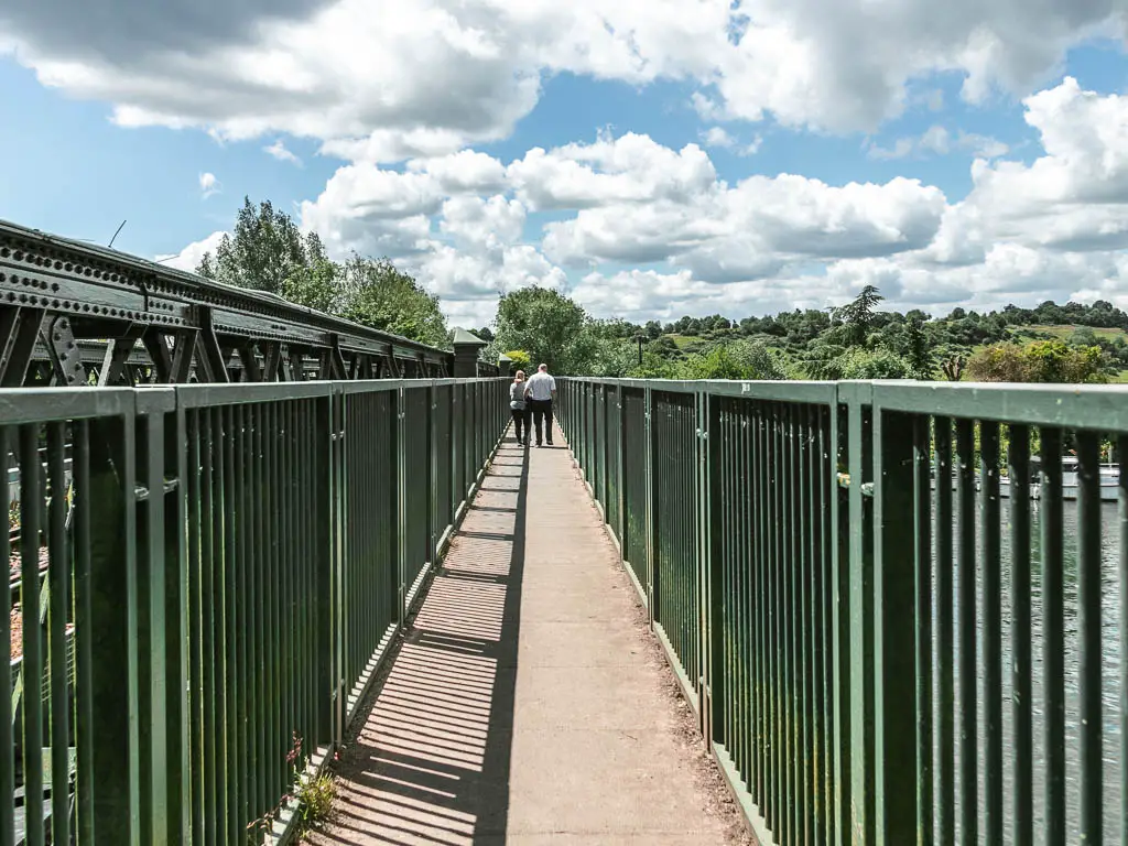 Looking along the bridge leading straight ahead, lined with metal railings. There are two people walking along it ahead.