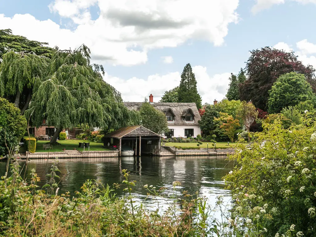 Looking over the bushes to a white walked thatched roofed cottage on the green on the other side, on the walk between Marlow and Maidenhead. There is a wooden boat garage in front of the cottage.