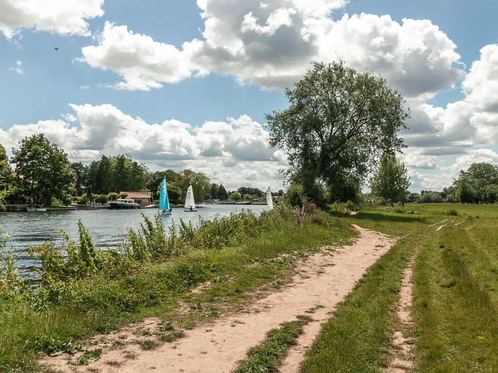 A dirt trail leading ahead through the grass field, with the river to the left and a few small sailing boats in the river.