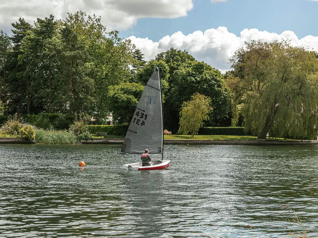 A small sailing boat on the river, on the walk from Marlow to Maidenhead. There is a person in the boat. There is a green with trees on the other side of the river.