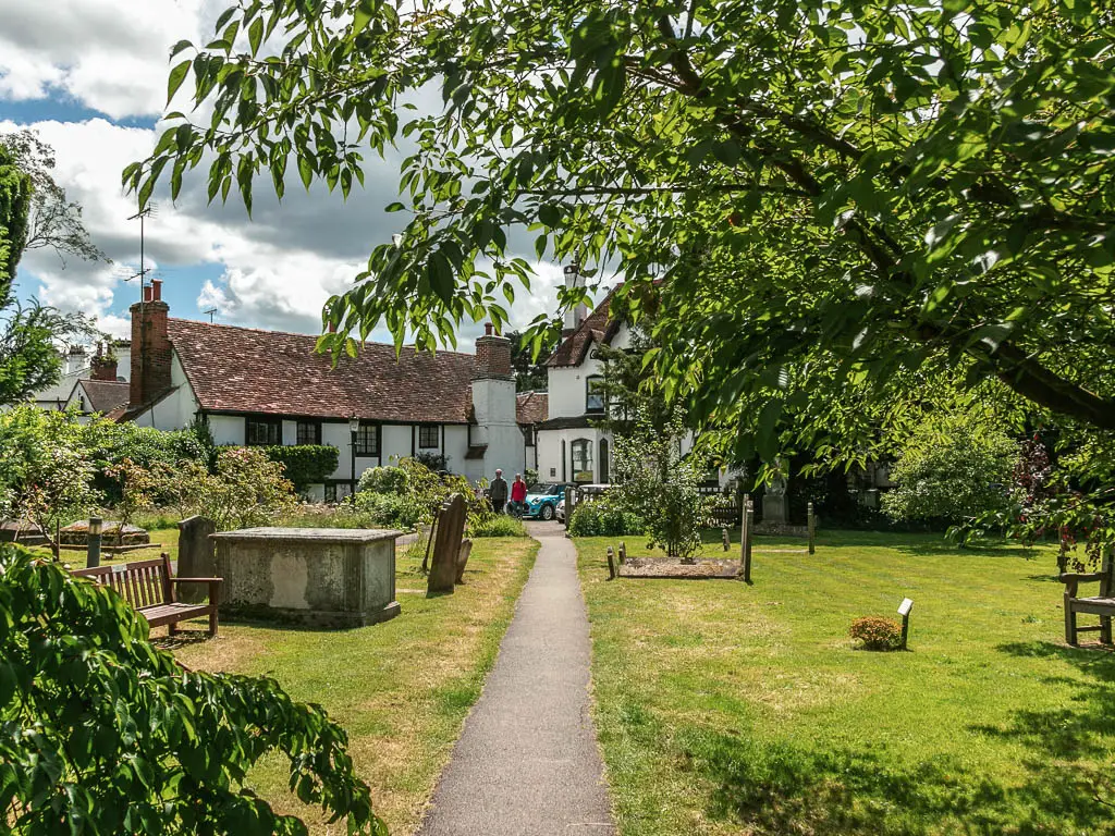 A narrow straight path leading through the neatly cute grass of the graveyard with some white walled buildings ahead. There are tree branches hanging into the frame from the right.