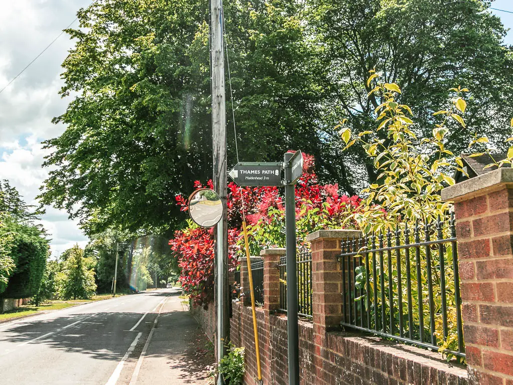 A thames path sign on the pavement pointing left across the road. There is a trick wall with metal railings to the right. There are bushes with green and red leaves behind the railings.