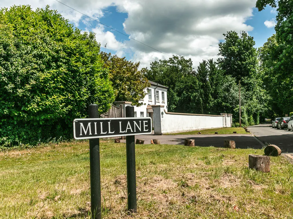 A road sign saying 'mill lane' in the grass front lawn. There is a white walled house behind partially hidden by big leafy trees and a white wall.