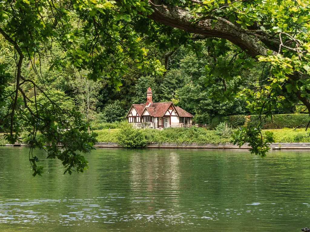 Looking across the river to a Tudor style cottage on the other side, on the walk from Marlow to Maidenhead. There are tree branches with green leaves hanging into the frame from above. There is a mass of green leafy trees behind the cottage.